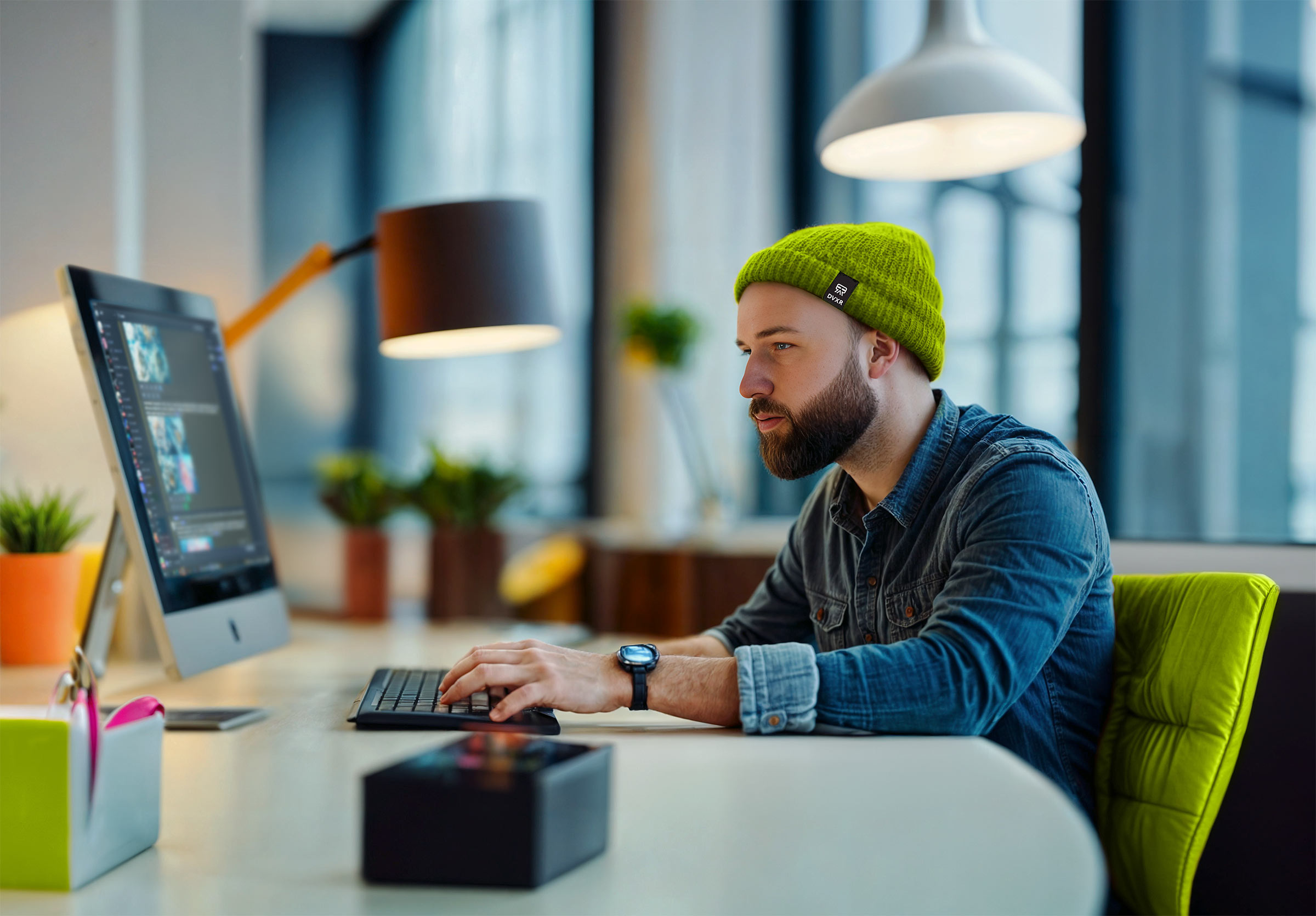 A young man in a green beanie working on a computer in a modern office.